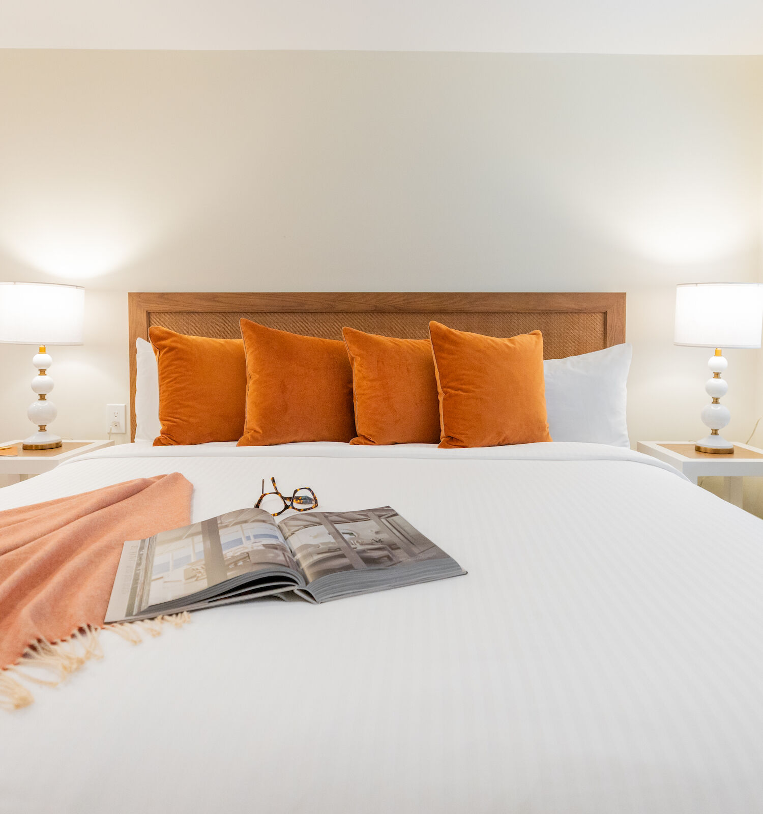 A neatly made bed with orange pillows and a matching throw blanket, a magazine and glasses on it, flanked by two lamps on bedside tables.