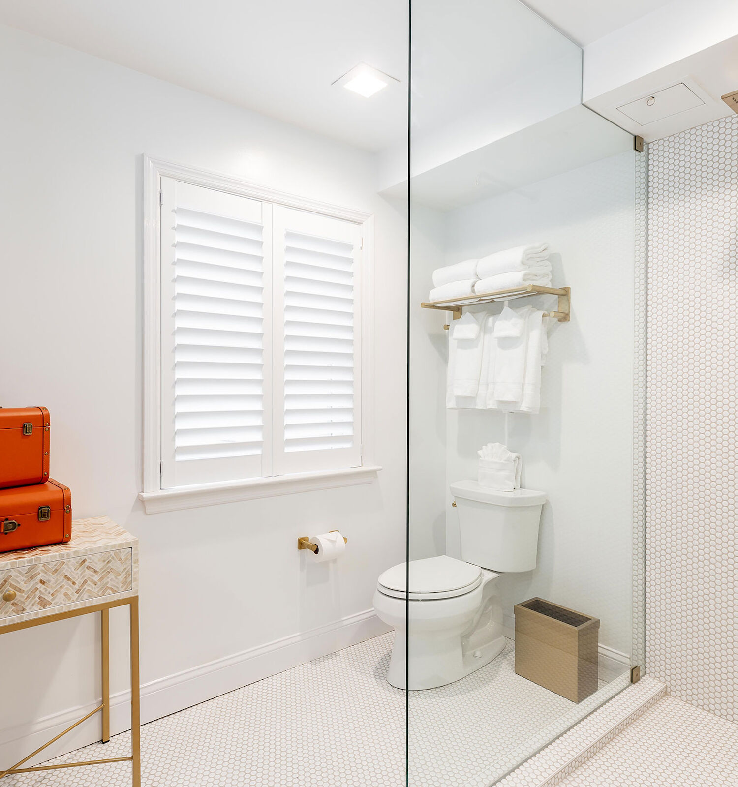 A modern bathroom with a shower, toilet, and sink. The decor includes orange suitcases on a table and white towels on a shelf.