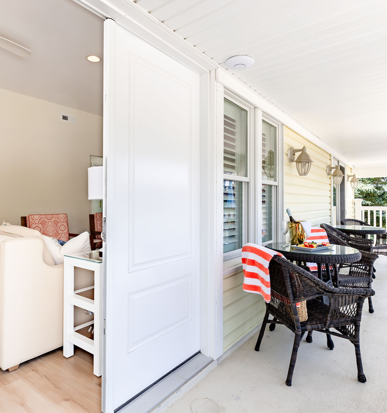 A cozy porch with wicker chairs, a red-striped towel, small tables, and windows, adjacent to a living room with a couch and a ceiling fan.