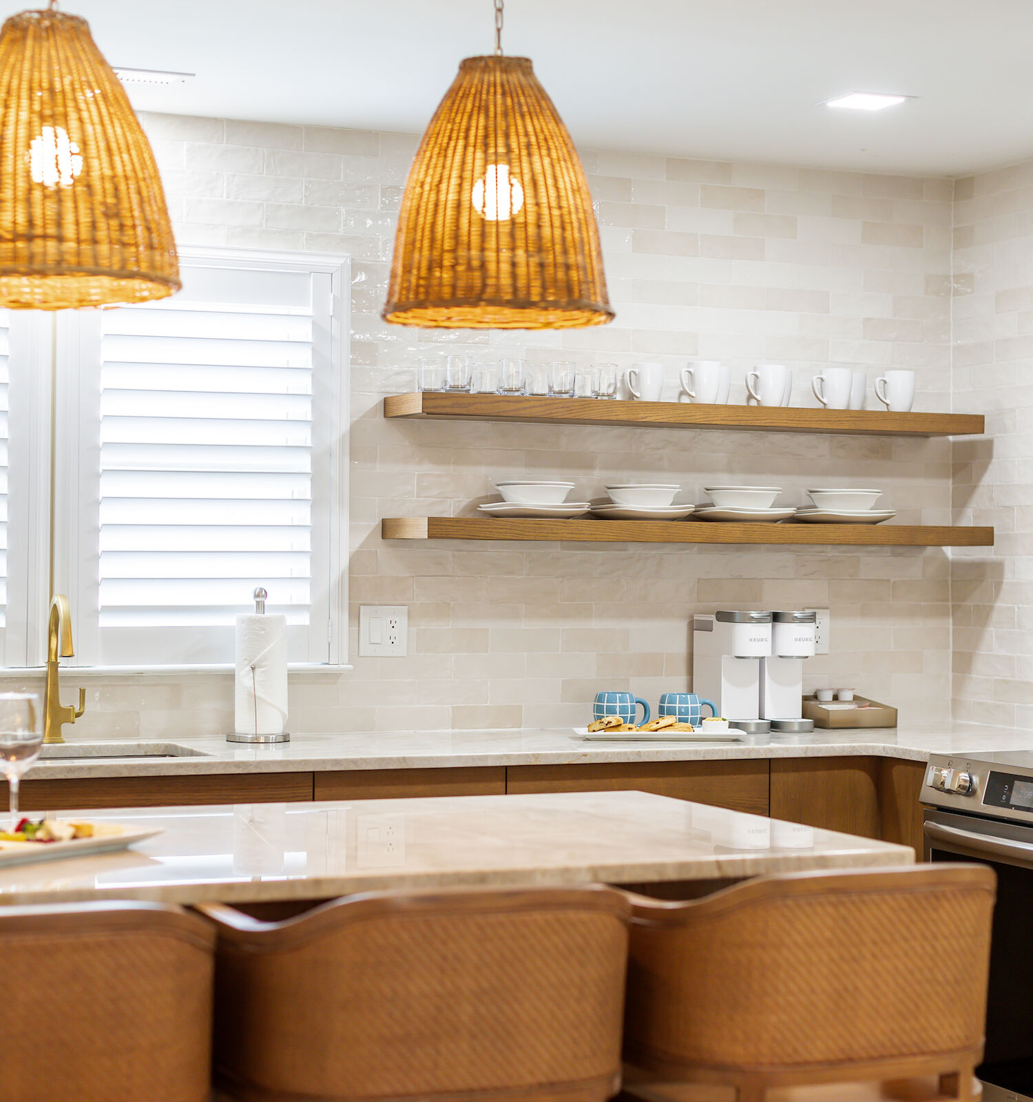 A modern kitchen with wooden cabinets, hanging wicker lights, open shelves with dishes, a wine bottle, glasses, and a window with shutters.