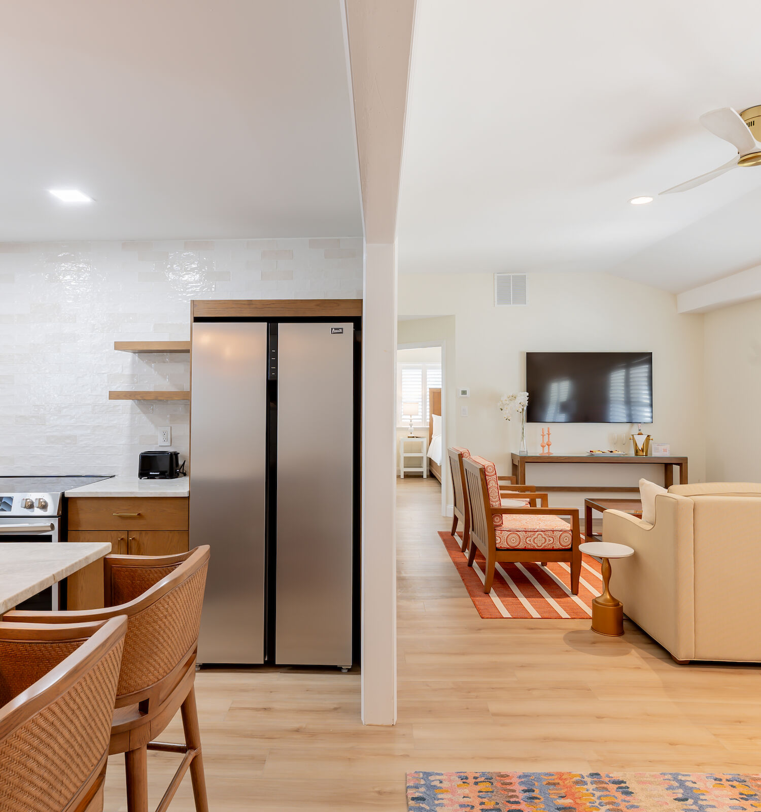 This image shows a modern open-concept kitchen and living room area with wooden furniture, a large refrigerator, and a TV mounted on the wall.