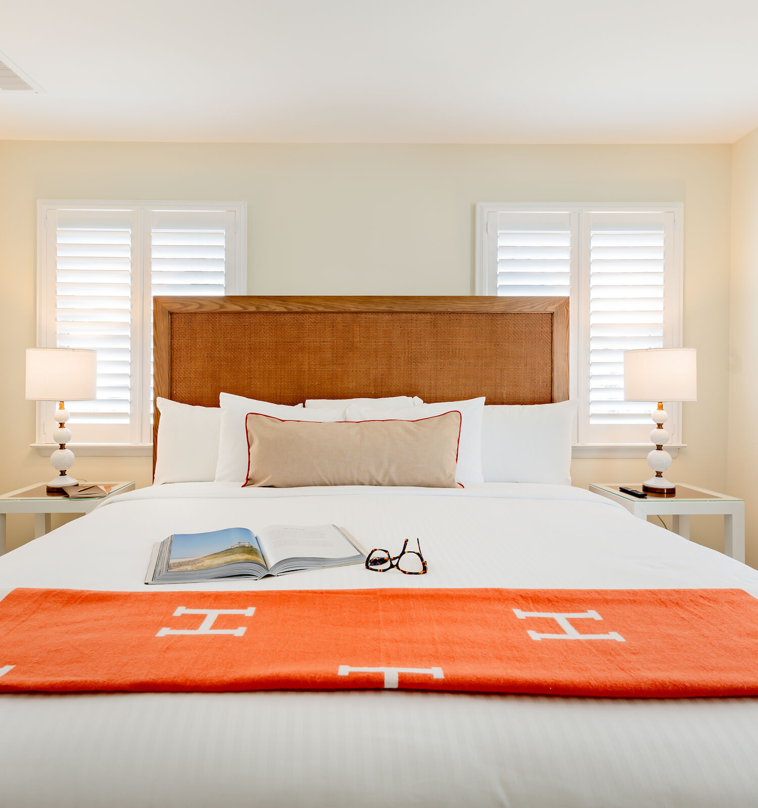 A cozy bedroom featuring a neatly made bed with orange blanket, pillows, book, and glasses. Two nightstands with lamps flank the bed.