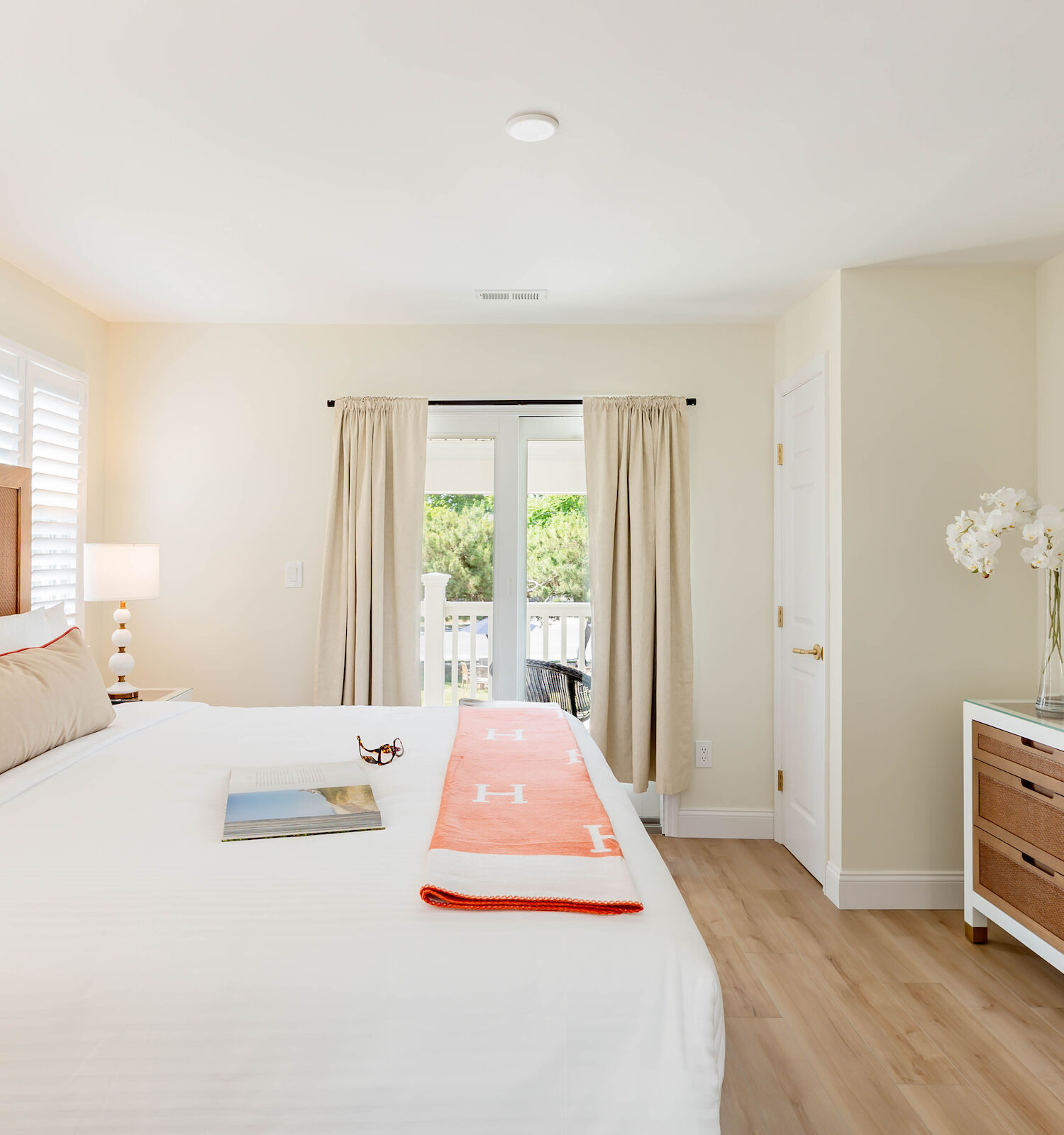 A modern bedroom with a large bed, a wall-mounted TV, a dresser, doors leading to a balcony, and a nightstand with a lamp and floral decor.