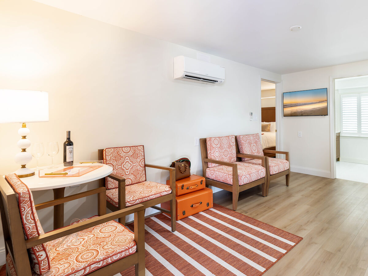 A cozy living area with patterned chairs, a round table with a bottle of wine, striped rug, wall-mounted air conditioner, and artwork on the wall.