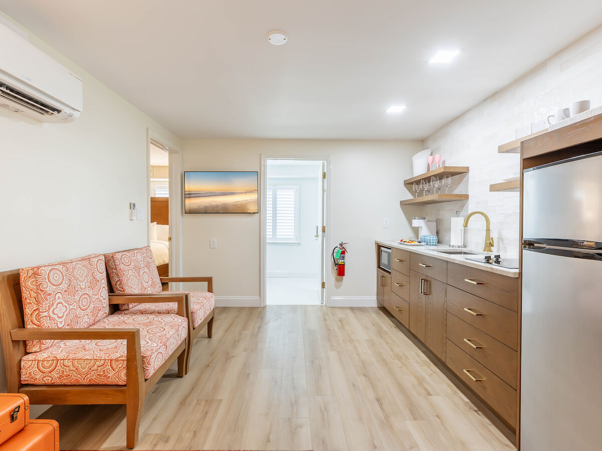 This image shows a modern room with two armchairs, a small kitchen area on the right, a door in the center, and a light wood floor.