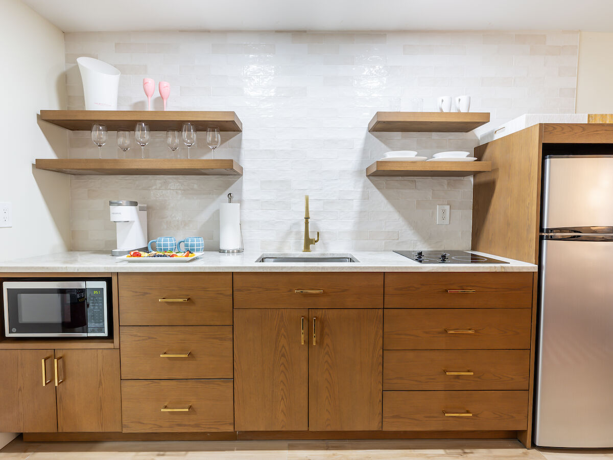 A modern kitchen with wooden cabinets, open shelves, white backsplash, microwave, sink, stovetop, and a stainless steel fridge and freezer.