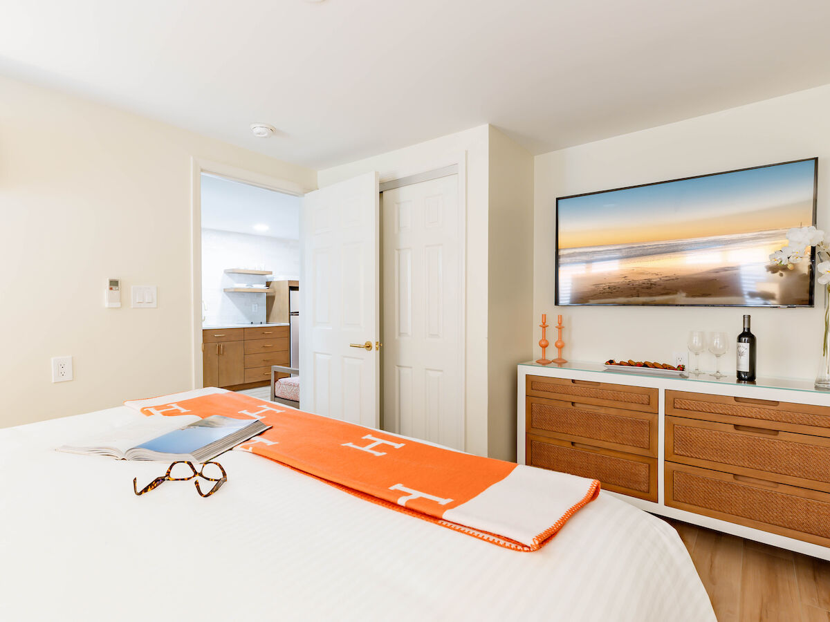 A cozy bedroom with a bed, an orange blanket, a pair of glasses, a TV, and a dresser with decor items including a wine bottle and glasses.