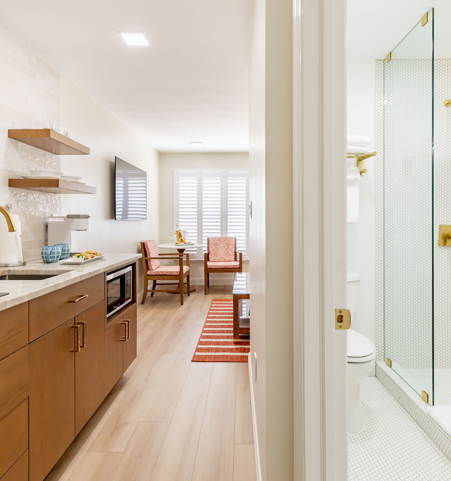 A modern, compact studio apartment featuring a kitchenette with wooden cabinetry and a bathroom with a glass-enclosed shower on the right side.