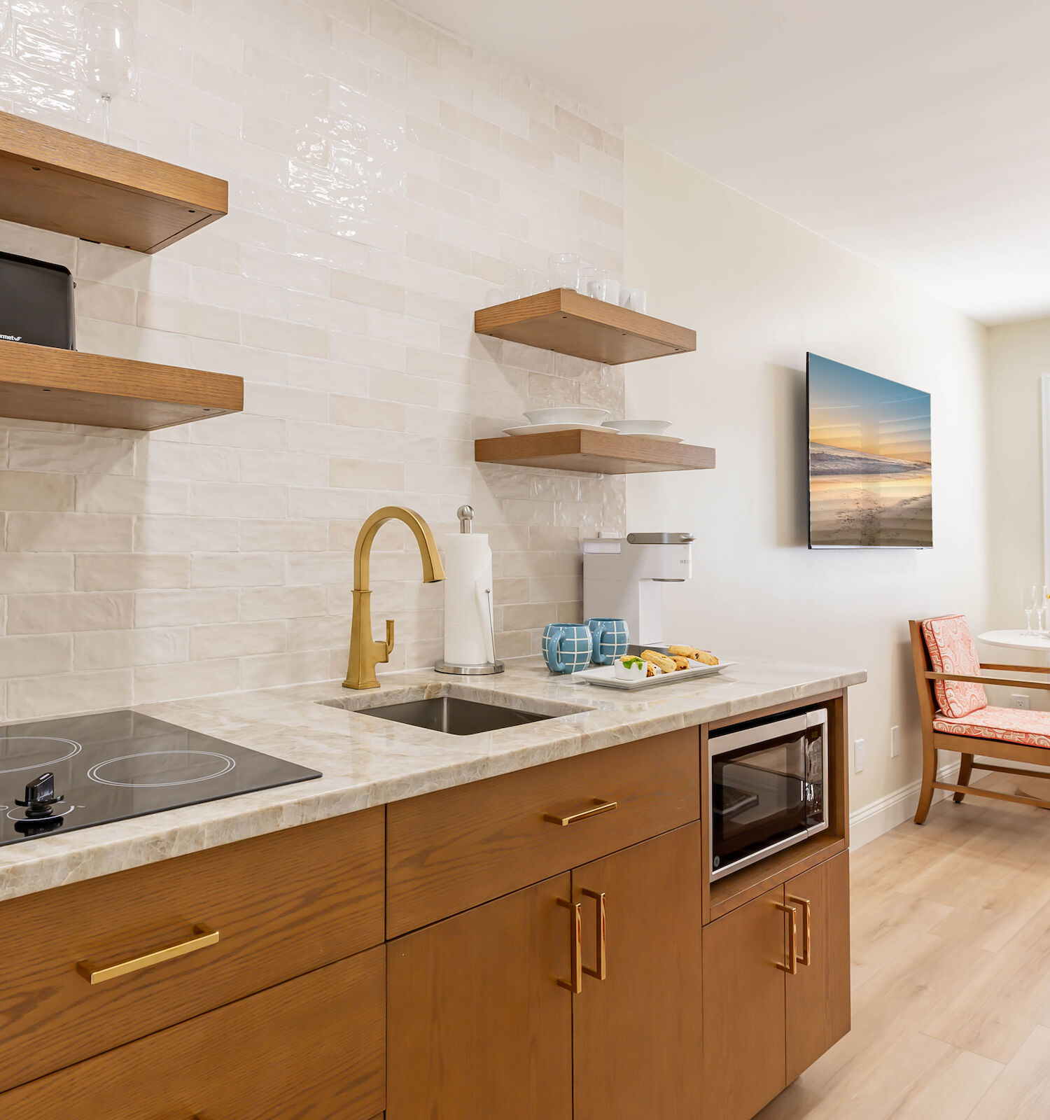 A modern kitchen with a stove, sink, microwave, and shelving. A small dining area with table, chairs, and window with white shutters is visible.