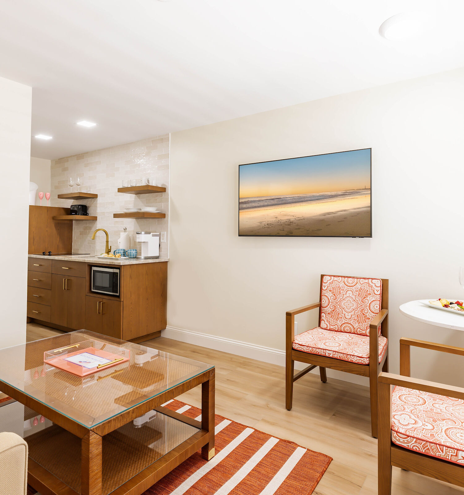 A cozy, modern living area with a small kitchen, a wall-mounted TV, two chairs, a sofa, and a coffee table. It features a minimalist design.