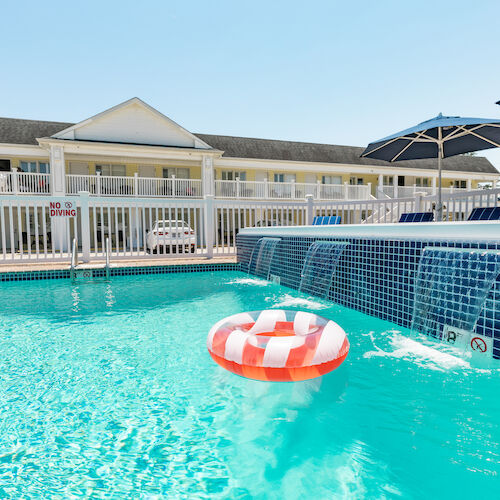 A swimming pool with a striped inflatable ring, adjacent to a building with rooms and a public space under umbrellas on a sunny day.