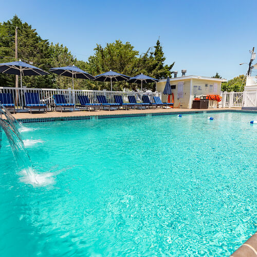 A clean swimming pool with cascading water, lounge chairs, and umbrella-shaded tables in a sunny outdoor area near a building with a white fence.