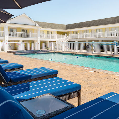 A courtyard with a swimming pool surrounded by blue cushioned lounge chairs and an umbrella, within a two-story building complex.