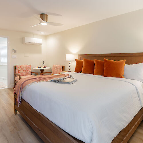 A modern bedroom with a large bed, orange pillows, a bedside table with lamps, a seating area, a ceiling fan, and a view into the bathroom.