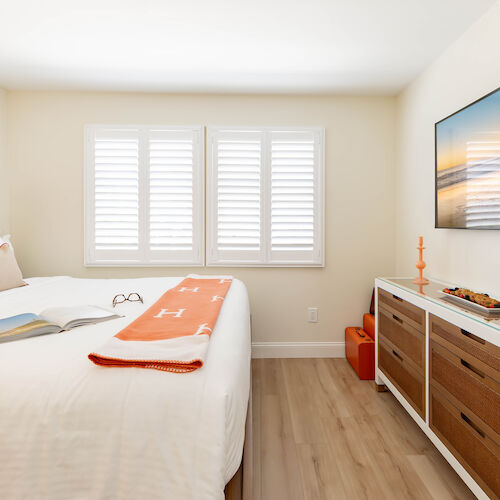 A cozy bedroom with a neatly made bed, a dresser with a TV above, books, glasses, a candle, wine bottle, and glass. Bright with large windows ending the sentence.