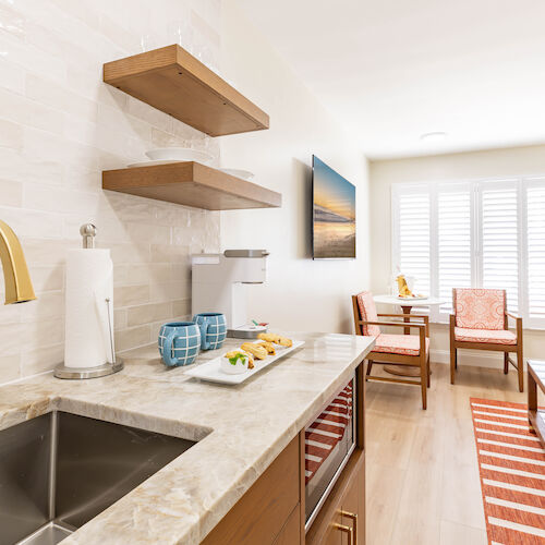 The image shows a modern kitchen with a sink, open shelves, and a countertop. A dining area with chairs and a TV mounted on the wall can be seen.