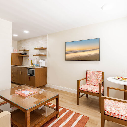 A cozy living area with a kitchenette, a wall-mounted TV, a coffee table, two chairs, a small round table, and minimal decor.