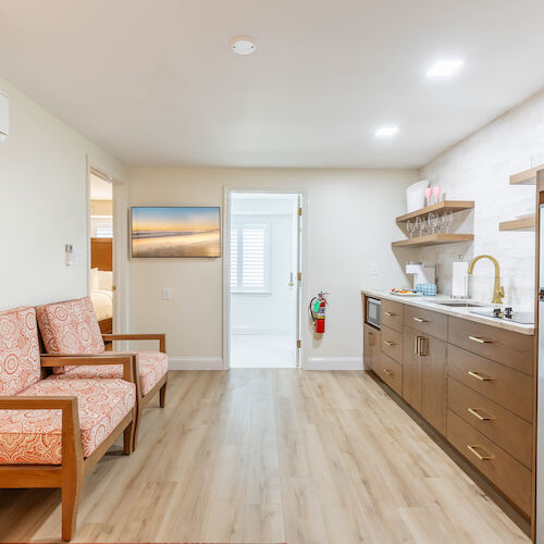 A well-lit room with a small kitchenette, two patterned chairs, a wooden floor, and an air conditioner on the wall. The room appears clean and modern.