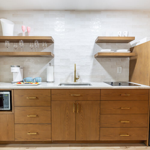 A tidy kitchen with wooden cabinets, open shelves holding dishes and glasses, a microwave, sink, and stainless steel refrigerator ending the sentence.