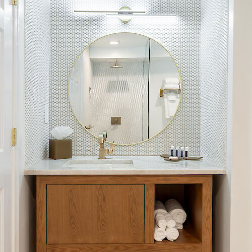 This image features a bathroom vanity with a round mirror above it, a sink, a neatly folded stack of towels, and small toiletries on the countertop.