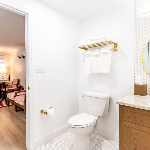 A bathroom with a toilet, a vanity with a round mirror, and towels on a gold rack. There's an adjoining room with a seating area and a striped rug.