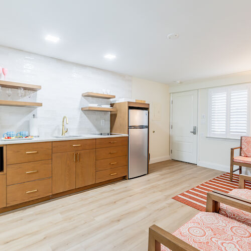 A modern kitchen and living area with wooden cabinets, a microwave, a fridge, and seating on a patterned rug.