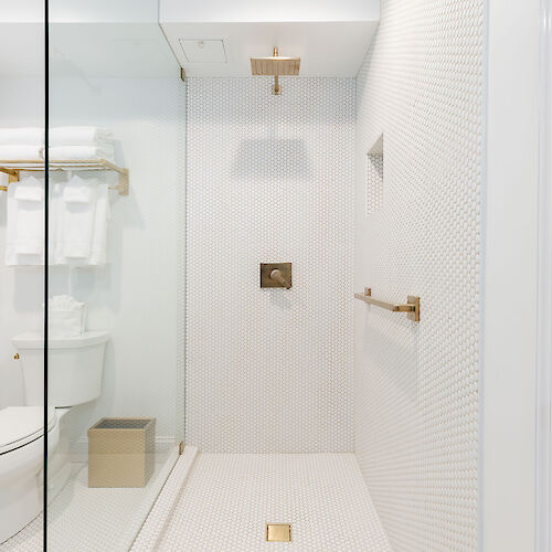 A modern, white-tiled bathroom with a glass shower enclosure, golden fixtures, a toilet, and a wooden basket is shown in the image.