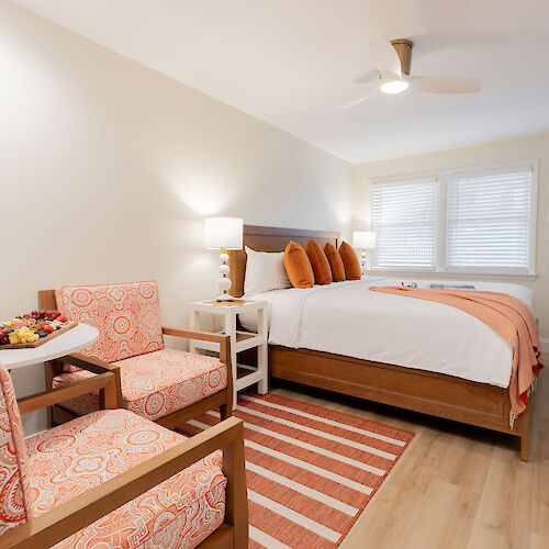 A cozy bedroom with a neatly made bed, orange and white decor, patterned armchairs, a striped rug, and modern bedside lamps and windows.