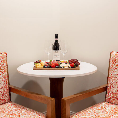 A small table with a bottle of wine, two glasses, and a tray of assorted foods is flanked by two patterned chairs.