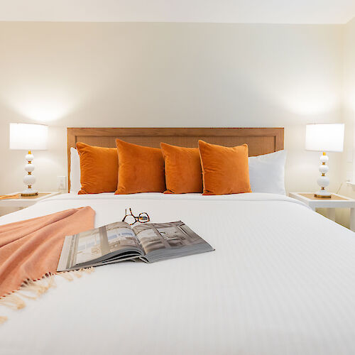 A neatly made bed with orange pillows, a book and glasses, a blanket draped over the bed; two bedside lamps and a window with blinds.