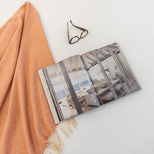 An open magazine showing modern interiors, a pair of glasses, and a peach-colored blanket are placed on a white surface.