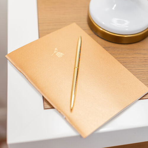 A gold-colored notebook and pen sit on a white and wooden nightstand, next to a white lamp with gold accents.