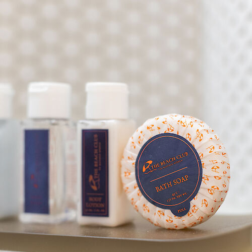 The image shows three small bottles and a wrapped soap bar on a tray, likely hotel toiletries, with a patterned background.