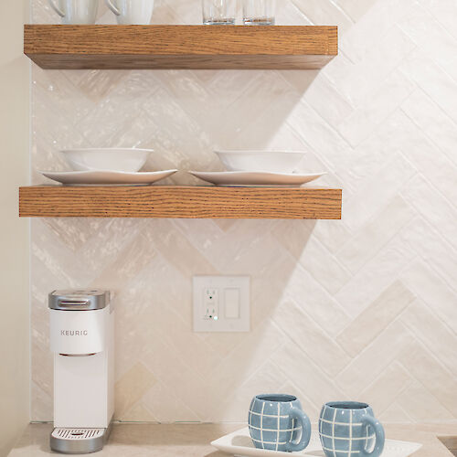 A kitchen scene with two wooden shelves holding mugs, glasses, and plates. Blue checkered mugs on a tray sit on the counter with a water filter nearby.