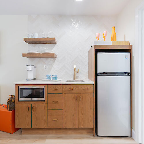 A compact kitchen setup with wooden cabinets, a countertop with a small sink, microwave, mini fridge, shelves, and a door to the right ending the sentence.