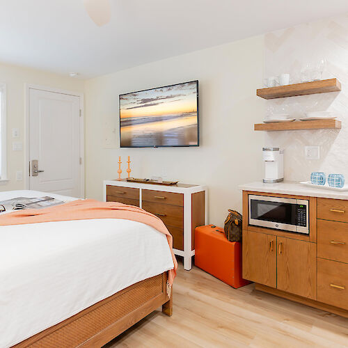 This image shows a modern bedroom with a bed, wall-mounted TV, wooden furniture, and a small kitchenette area with a microwave and shelves, ending the sentence.