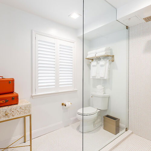 A modern bathroom with a walk-in shower, toilet, window, red suitcase on a table, towel rack holding white towels, and a waste bin next to the toilet.