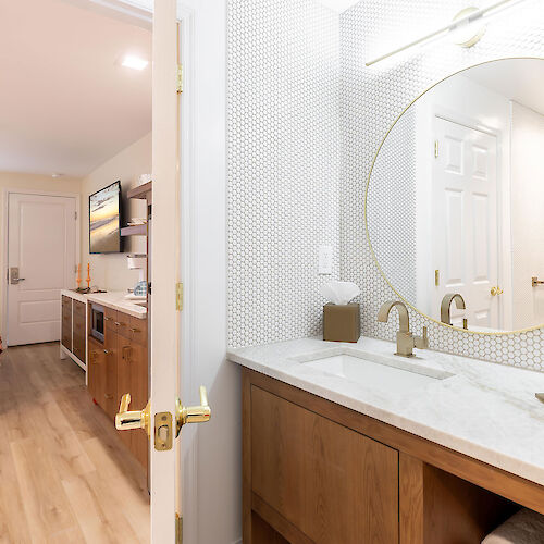 This image shows a modern hotel room with a bathroom visible from the open door. The bathroom has a round mirror, a sink, and toiletries.