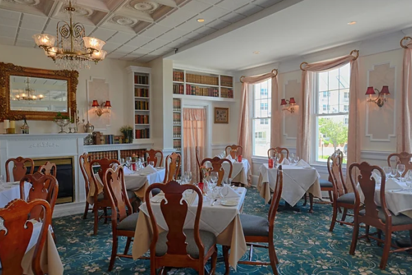 A beautifully decorated dining room with elegant tables, chairs, chandeliers, large windows, and a fireplace with a mirror above it is shown.