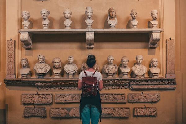 A person with a backpack is viewing a display of various classical busts and stone carvings in a museum setting, standing in front of a beige wall.