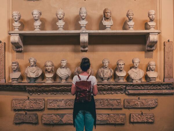A person with a backpack is viewing a display of various classical busts and stone carvings in a museum setting, standing in front of a beige wall.