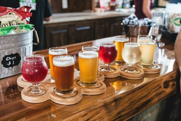 A wooden bar counter displays an array of eight colorful beer samples in small glasses, arranged on wooden trays shaped like paddles.