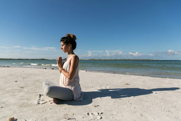 A person is meditating while seated on a sandy beach near the ocean, under a clear blue sky, surrounded by calm and peaceful scenery.