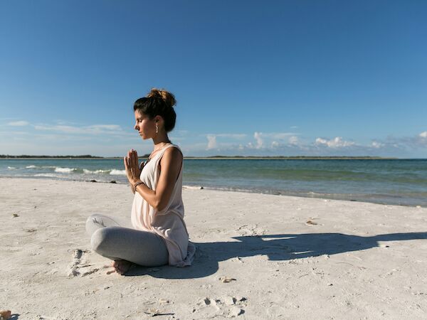 A person is meditating while seated on a sandy beach near the ocean, under a clear blue sky, surrounded by calm and peaceful scenery.