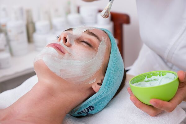 A person is receiving a facial treatment with a mask being applied on their face by a professional using a brush, holding a bowl of cream.