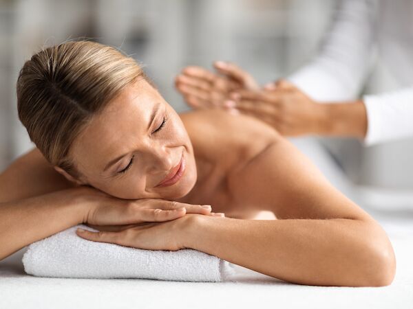 A woman is receiving a massage, lying on a white towel, looking relaxed with eyes closed while a therapist massages her back.