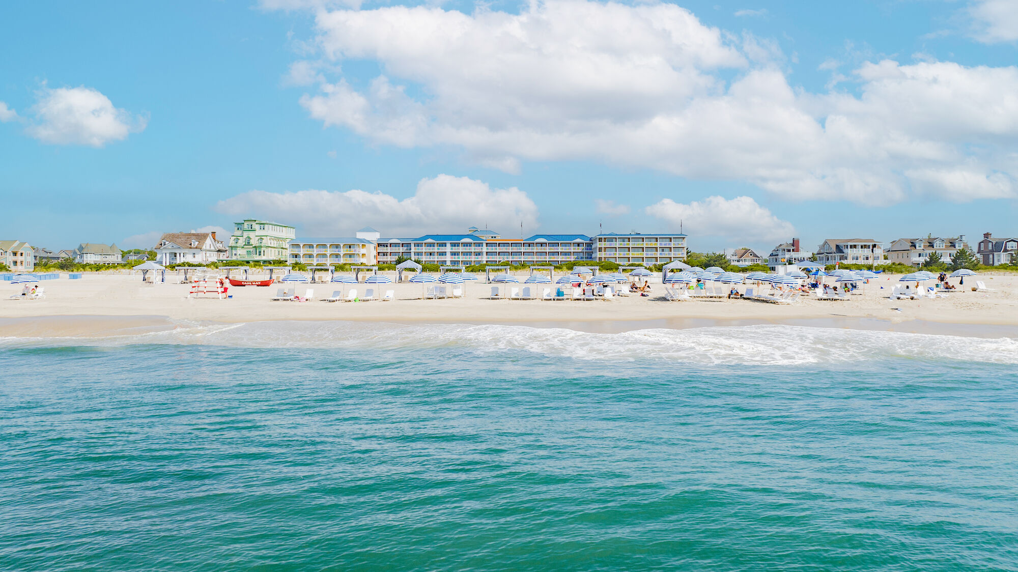 A scenic beach with clear blue water, white sand, beachgoers, umbrellas, and buildings in the background under a bright, partly cloudy sky.