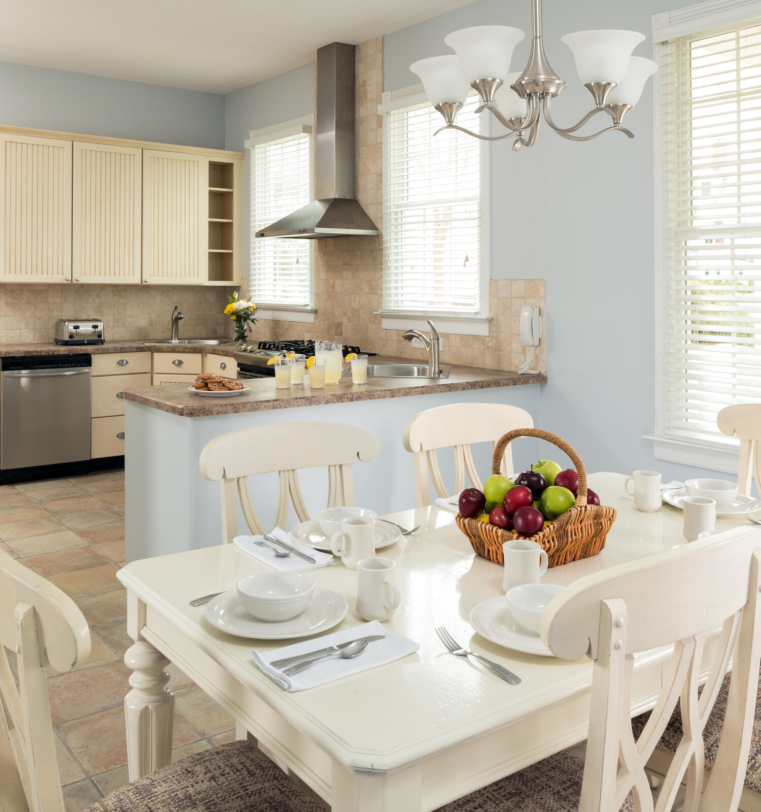 A kitchen and dining area with a table set for four, including a fruit basket, white cabinetry, stainless steel appliances, and a chandelier.