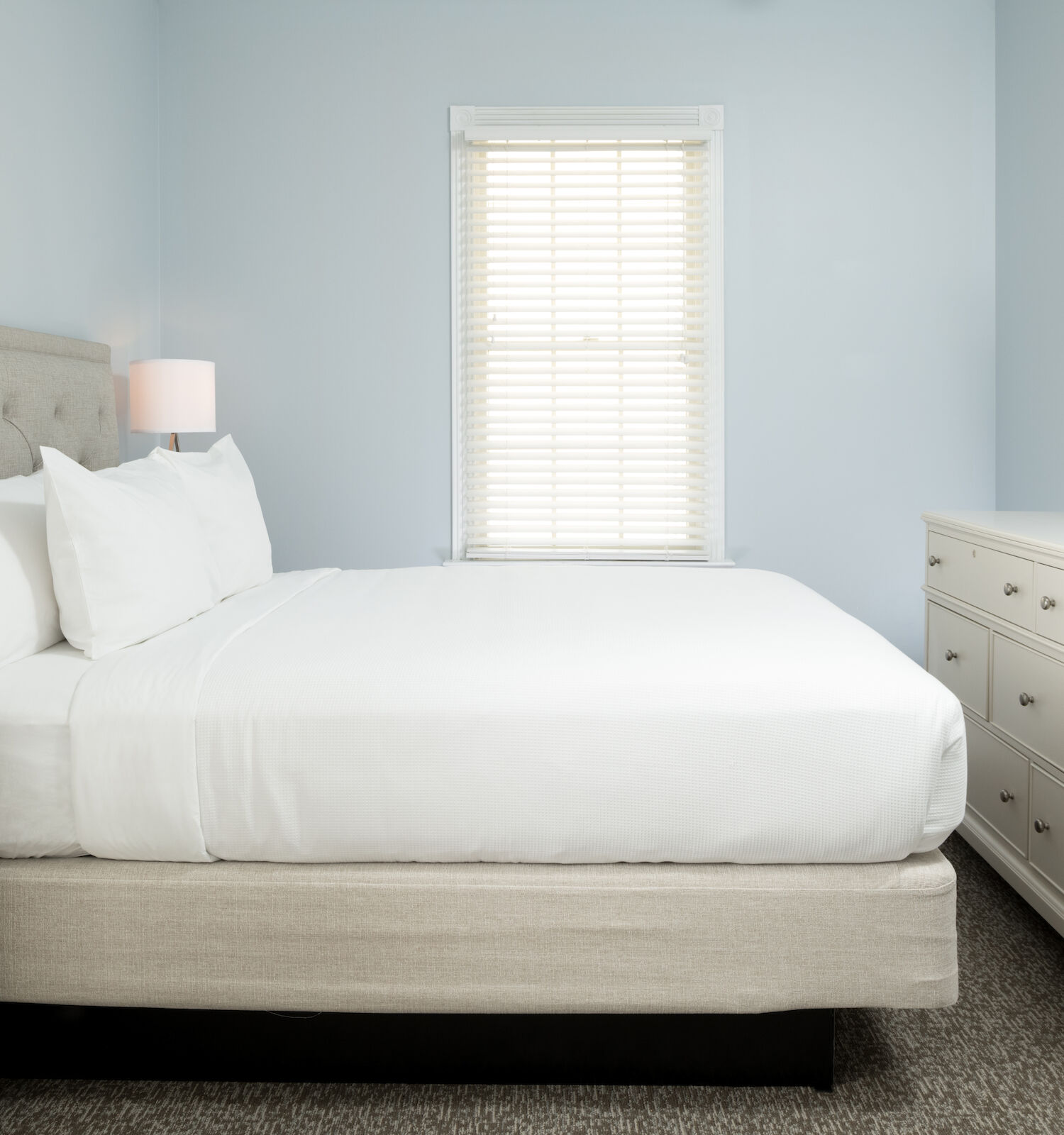 This image shows a cozy bedroom with a neatly made bed, two nightstands with lamps, a dresser, a TV mounted on the wall, and a window with blinds.