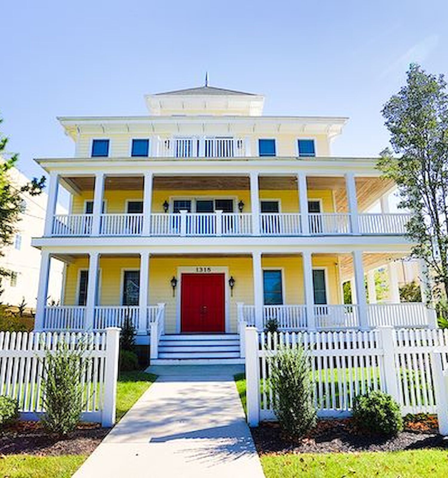 A large yellow beach house with multiple balconies, a red door, and a white picket fence. A sign reads 